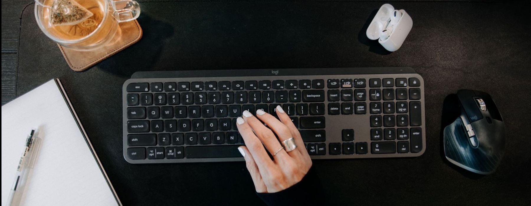 woman's hand on a keyboard surrounded by office items and a cup of tea on a coaster